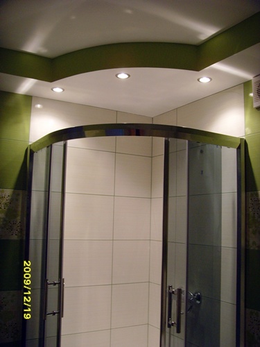 Wykończona łazienka z kabiną prysznicową narożną z chromowanymi detalami, białe płytki i zielone akcenty kolorystyczne na ścianach i suficie, oświetlenie punktowe.